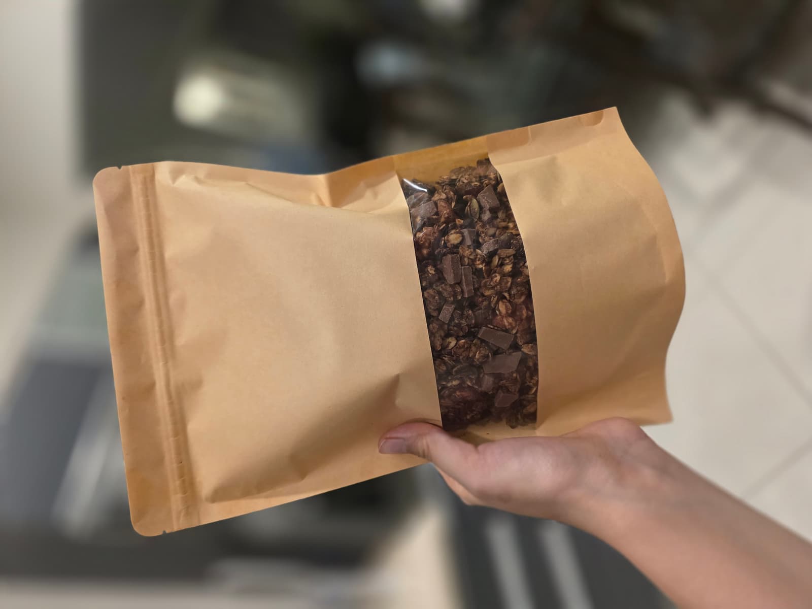 Hand holding a kraft pouch of NutriHome chocolate granola, chocolate chunks visible through the window